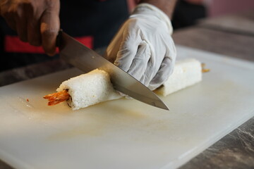 Japanese sushi is cutted and plated by gloves wearing hand chef with sharp knife, shrimp and sauces are visible in between sticky rice and bamboo role and firing the sushi role with burner 