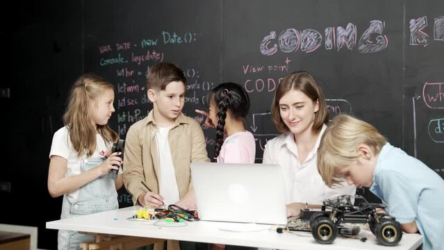 Caucasian teacher using laptop and explain about engineering system while diverse student holding electronic equipment in front of blackboard with prompt or code written in STEM class. Erudition.