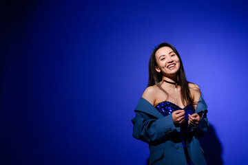 Cheerful and stylish asian woman in dress and jacket posing on blue background 