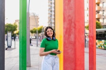 A vibrant urban scene captures a smiling woman with her smartphone and headphones, leaning against...