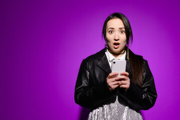 Shocked asian woman in leather jacket holding smartphone and looking at camera on purple 