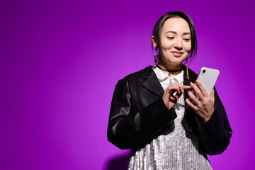 Smiling young asian woman in dress with sequins using smartphone on purple 