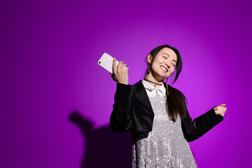 Smiling asian woman in stylish party dress holding smartphone and dancing on purple background 