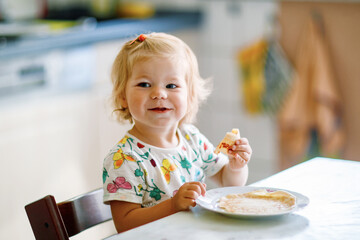 Happy little toddler girl eating delicious pancakes sitting in the kitchen. Cute child tasting...