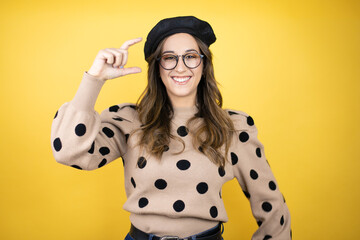 Young beautiful brunette woman wearing french beret and glasses over yellow background smiling and...