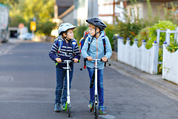 Two school kid boys in safety helmet riding with scooter in the city with backpack on sunny day....