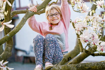 Cute spring fashion preschool girl with glasses under blossom magnolia tree. Little happy child and...