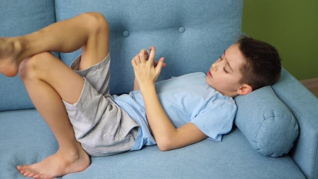 A little teenage boy lies in the room, engrossed in his smartphone. Children and gadgets. Online learning, games, children apps.
