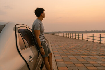 Asian man standing, relaxing and leaning the car, looking out the river at sunset time, thinking...