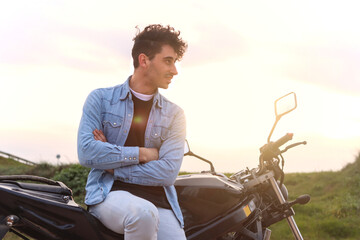Curly haired Caucasian young man sitting on one side of his motorbike with crossed arms looking...