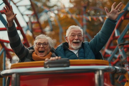 Hombre y mujer senior de m&aacute;s de 70 a&ntilde;os con gafas graduadas, sentados y felices , levantando los brazos, en una monta&ntilde;a rusa de un parque de atracciones sobre fondo desenfocado 