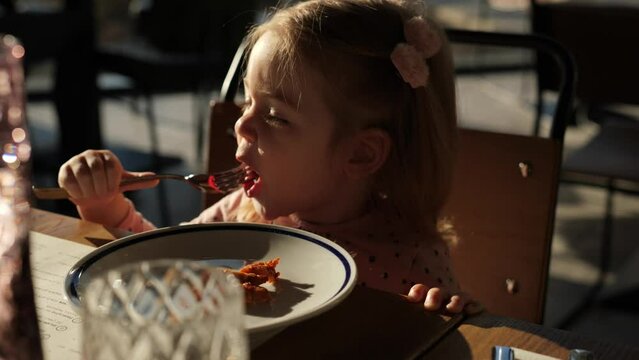 Adorable little girl eating meal using fork at the restaurant