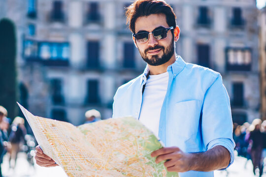 Half length portrait of bearded youg tourist in stylish sunglasses looking at camera while holding map in hands to find right direction of showplaces standing in downtown of architectural city