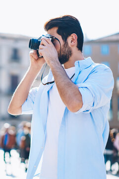 Skilled male amateur of making poto with vintage camera during trip.Hipster guy focusing to take pictures standing in urban setting.Photographer dressed in casual wear spending time outdoors on hobby