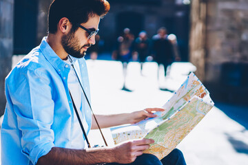 Side view of stylish casual dressed male bearded tourist in sunglasses looking at map in searching...