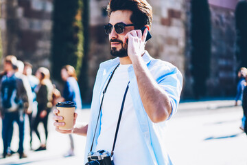 Stylish tourist in sunglasses talking on phone strolling with coffee to in architectural...