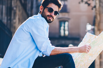 Portrait of cheerful bearded tourist in sunglasses sitting outdiirs on street of architectural city with old constructions while holding travel map for searching destination of showplaces