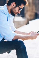 Obraz premium Side view of casual dressed male blogger in sunglasses typing text on smartphone fir publication on website sitting in downtown.Young man chatting with friends online via 4 internet connection