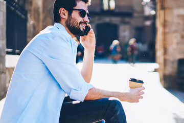 Bearded young man talking with operator about mobile benefits on smartphone sitting on street with...