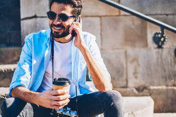 Cheerful young male photographer in stylish sunglasses talking with friends in roaming about good...