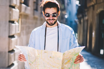 Cheerful bearded male lover of trips walking on streets of architectural town while searching right...