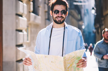 Half length portrait of happy casual dressed tourist in stylish sunglasses smiling at camera while strolling on narrow streets of old town holding travel map for searching right route of showplaces
