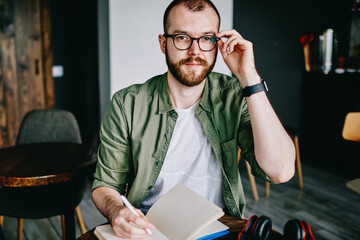 Young hipster male student in eyeglasses writing essay for homework bearded man thinking about tomorrow exam and looking aside, handsome creative author sitting at cafe interior with notebook