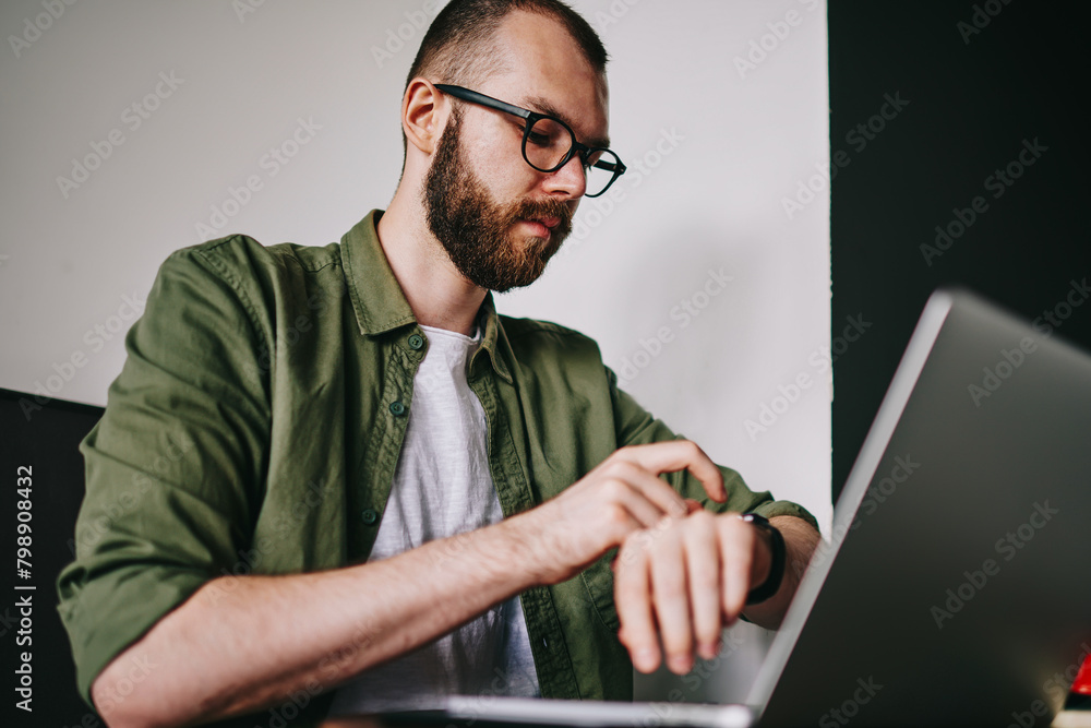 Poster young handsome man checking time on his smartwatch while chatting via laptop at favourite cafe, phot