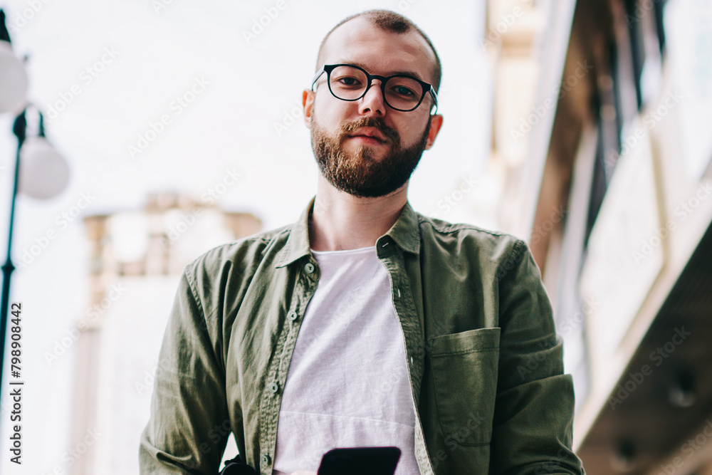 Wall mural portrait of young serious caucasian man in eyeglasses looking in camera while waiting friend on stre