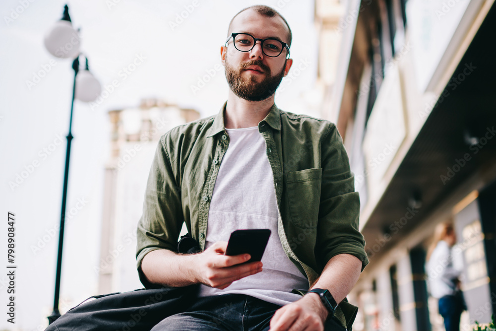 Poster portrait of young serious caucasian man in eyeglasses looking in camera while waiting friend on stre
