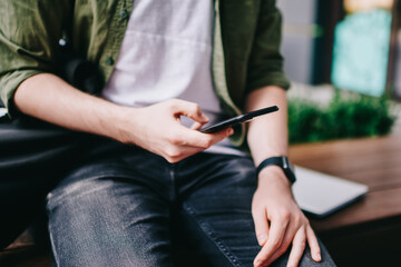 Cropped photo of hipster guy sitting outside and trying check mail by smartphone, male chatting with friends using new modern mobile phone while watching video online and reading notification