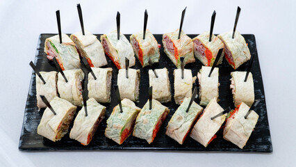 Different types of sushi rolls are neatly arranged on a black plate and strung on toothpicks. Catering