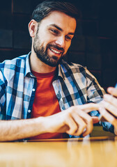Cheerful bearded young man dressed in casual outfit reading funny news in blog on smartphone device connected to 4G internet.Positive hipster guy watching video online on modern mobile phone