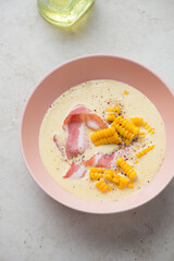 Corn chowder with bacon served in a roseate bowl, vertical shot on a light-beige stone background, high angle view