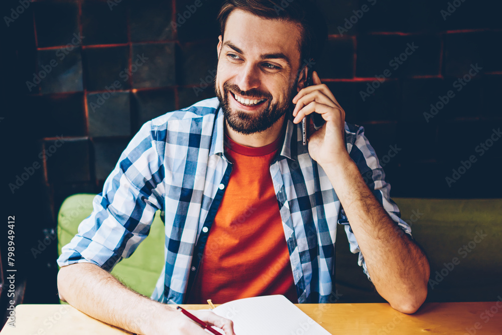 Poster successful bearded student calling on smartphone device while doing homework in notepad sitting at t