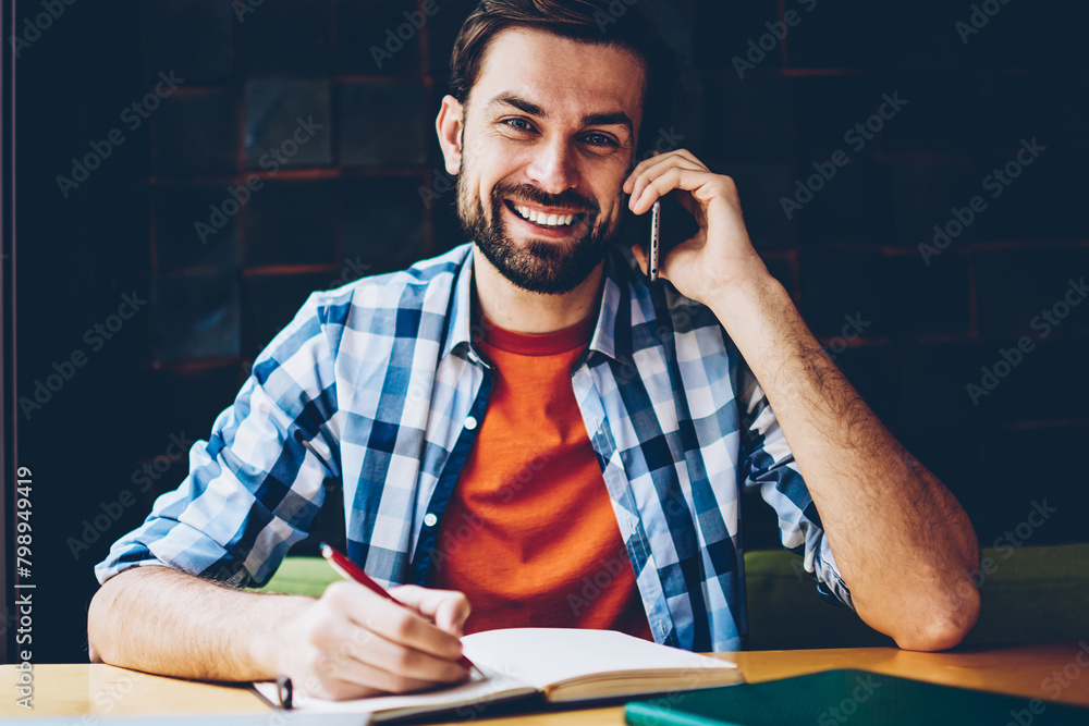Aufkleber portrait of successful bearded student smiling at camera while calling on smartphone and making note