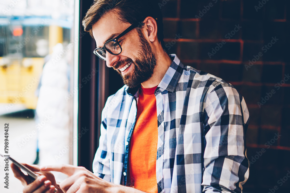 Poster joyful hipster young man in eyewear typing text message on smartphone device.positive student readin