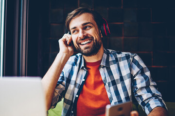 Positive bearded blogger enjoying electronic music in headphones connected via bluetooth to...