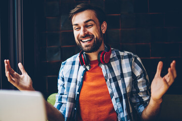 Cheerful male freelancer with stylish headphones on neck laughing during friendly conversation via...