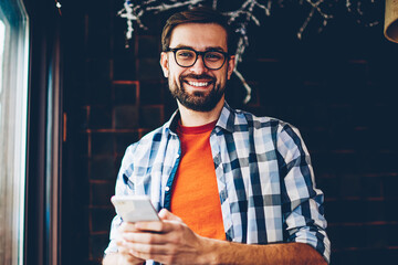 Portrait of happy bearded young man smiling at camera while typing text message on smartphone...