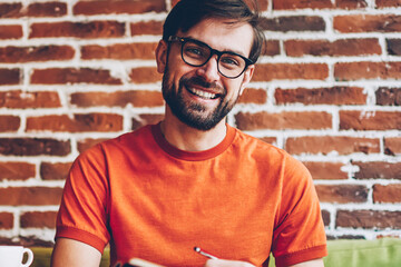 Half length portrait of successful bearded young man in eyewear smiling at camera while writing...