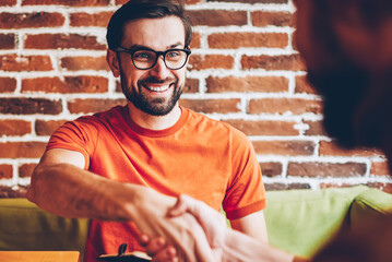 Cheerful bearded young man in eyeglasses handshaking with friend during meeting in coworking...