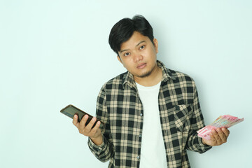 Young Asian man wearing brown flannel shirt showing confused face expression when holding mobile...