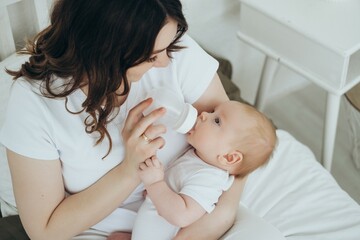 a mother feeds a baby from a bottle, a happy young mother feeds a newborn baby with milk or formula and smiles on the bed at home, a loving family holds their son in their arms. baby food