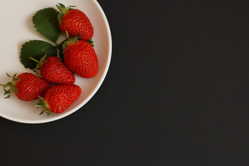 Red strawberries on white plate on black background top view.