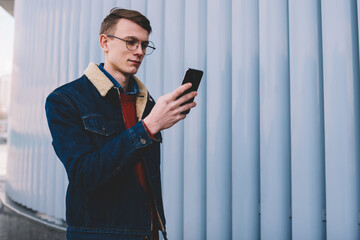 Concentrated young man dressed in stylish denim jacket watching videos on websites on smartphone...