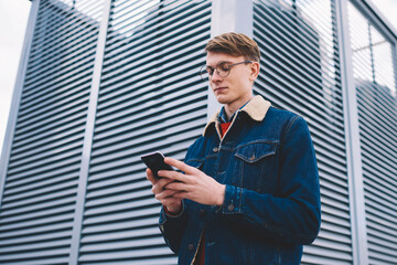 Young man in casual denim outfit reading notification on smartphone standing in downtown.Hipster guy watching video in blog on mobile phone via free 4G during walk in urban setting