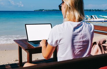 Back view of young blonde businesswoman working on ocean beach enjoying remote job in trip, hipster...