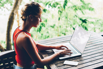 Skilled female blogger concentrated on keyboarding on netbook article for web page sitting at table...