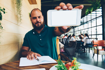 Portrait of serious bearded male food blogger holding smartphone for making picture of meal and...